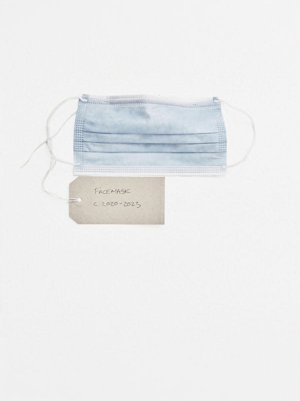 Still-life of a facemask with an archival tag. The name of the object and the dates 2020-2023 are written on it.
